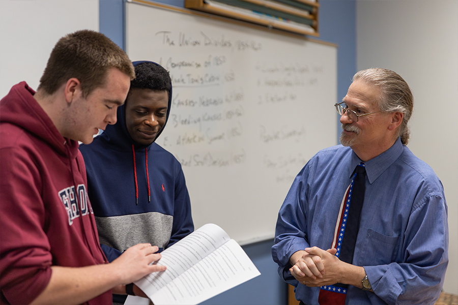 An instructor with graying hair, a goatee, and glasses stands smiling as he talks with two male students in a classroom. One student, wearing a maroon hoodie, holds open a textbook or packet that the group is reviewing together.
