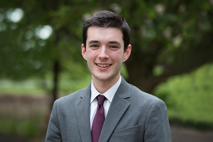 A young man in a gray suit and burgundy tie smiles outdoors with green trees in the background. A young man in a gray suit and burgundy tie smiles outdoors with green trees in the background.